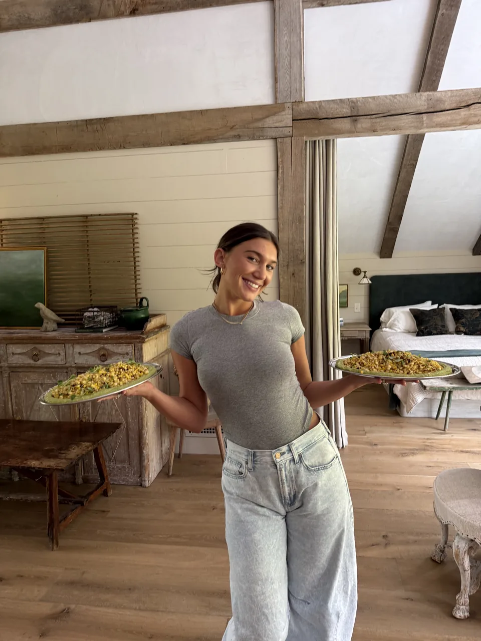 Chef Megan Luebke smiling and holding two platters of charred corn over a green herb puree in a farmhouse kitchen